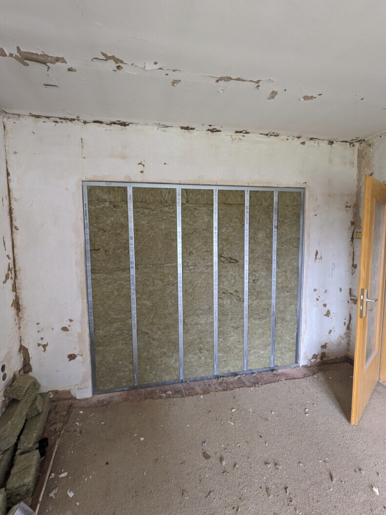 Raum im Umbau: freigelegte Wand mit Metallständerwerk und Dämmung, Tür offen.