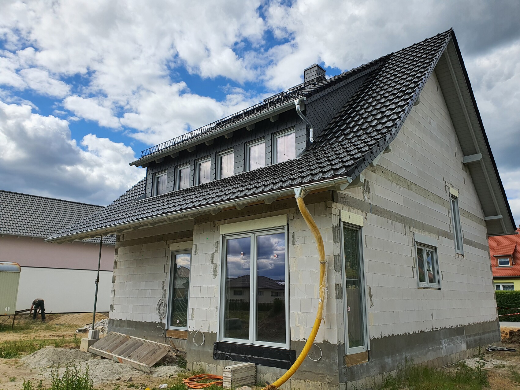 Zweistöckiges Haus im Rohbau mit grauen Dachziegeln; gelber Bau-Schlauch an der Fassade.