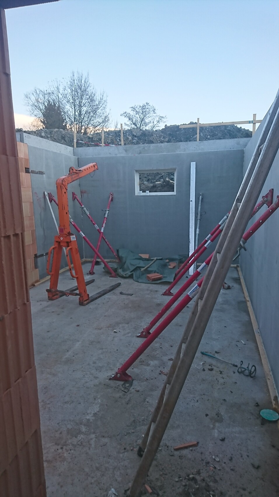 Baustelle: Betonraum mit Fensteröffnung, orange Stempel, rote Stützen, Baumaterial.
