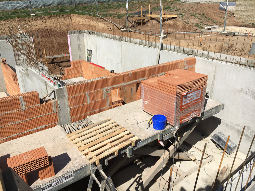 Rohbau-Baustelle: Ziegelwände entstehen, Paletten Ziegel, blauer Eimer auf Betonplattform.