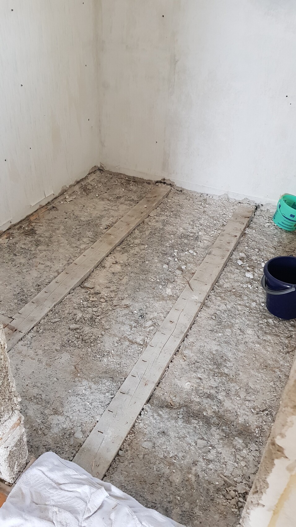 Bauzimmer mit freigelegtem Erdboden, zwei Holzlatten als Träger, Eimer und Bauabfälle.