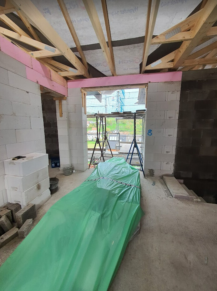 Rohbau-Innenraum: Holzdachkonstruktion, weiße Blöcke, grün abgedeckter Boden.