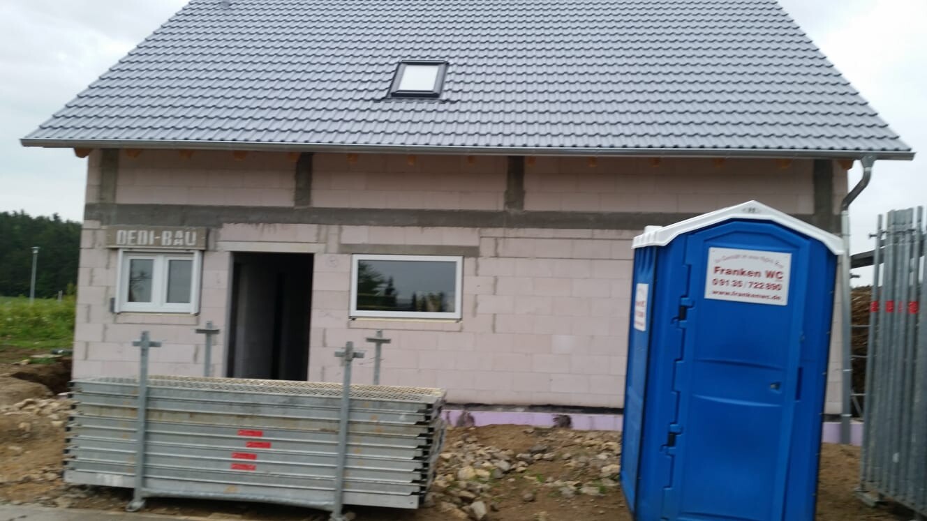 Halbfertiges Haus mit Ziegeldach, Bauzaun und blauem Porta-Potti