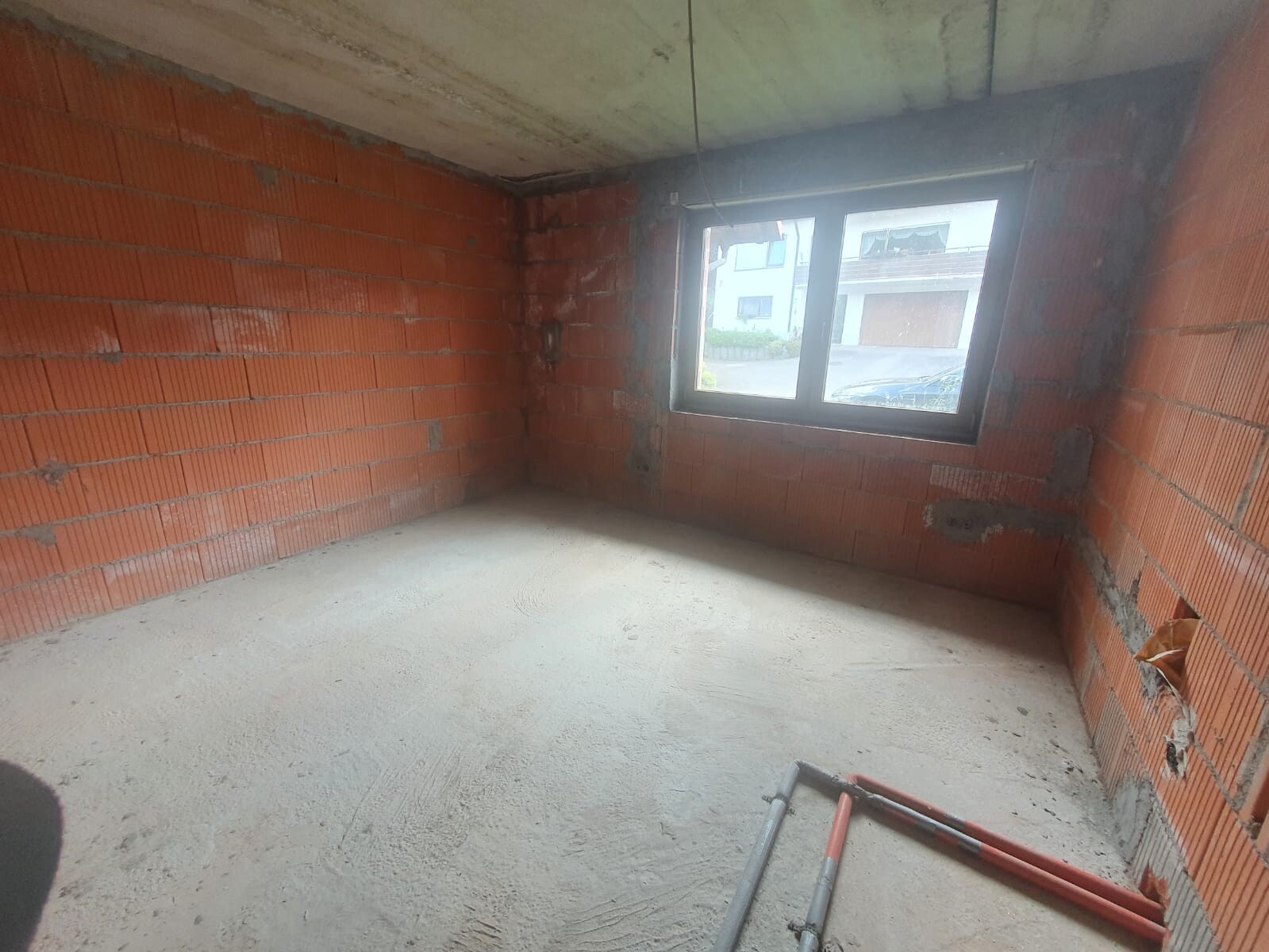 Unverputztes Bauzimmer mit roten Ziegelwänden, grobem Betonboden, Fenster und Rohren am Boden.