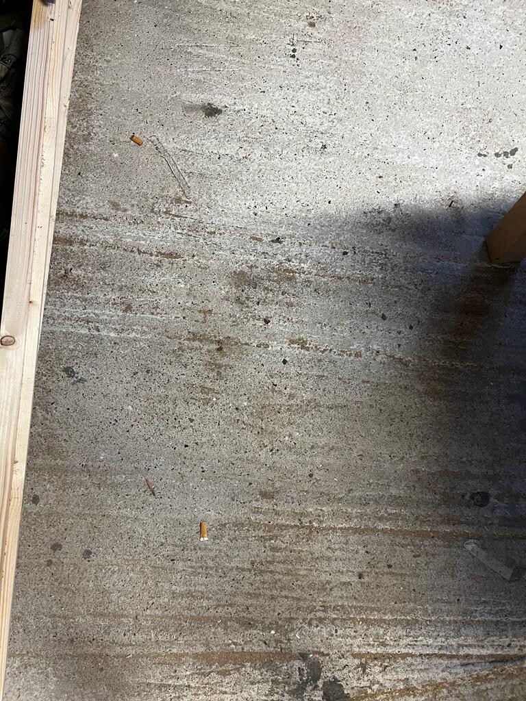 Grobe Betonbodenfläche mit Flecken, Staub und Zigarettenkippen; linker Holzbalken am Rand.