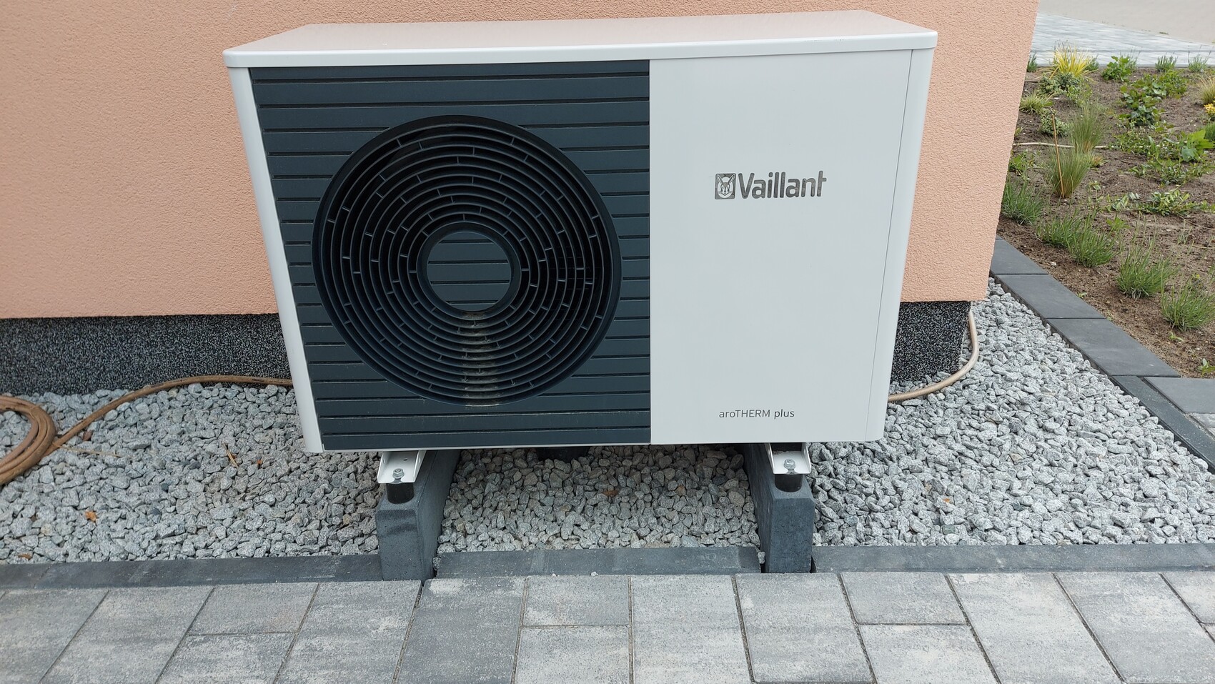 Außen-Luftwärmepumpe Vaillant aroTHERM plus mit großem Lüftergitter vor Kiesfläche neben Wand.