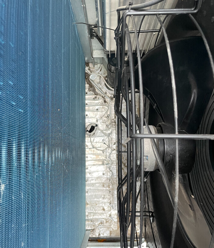Außenlüfter eines Klimageräts hinter Schutzgitter, links blaue Verkleidung, Kabel sichtbar.