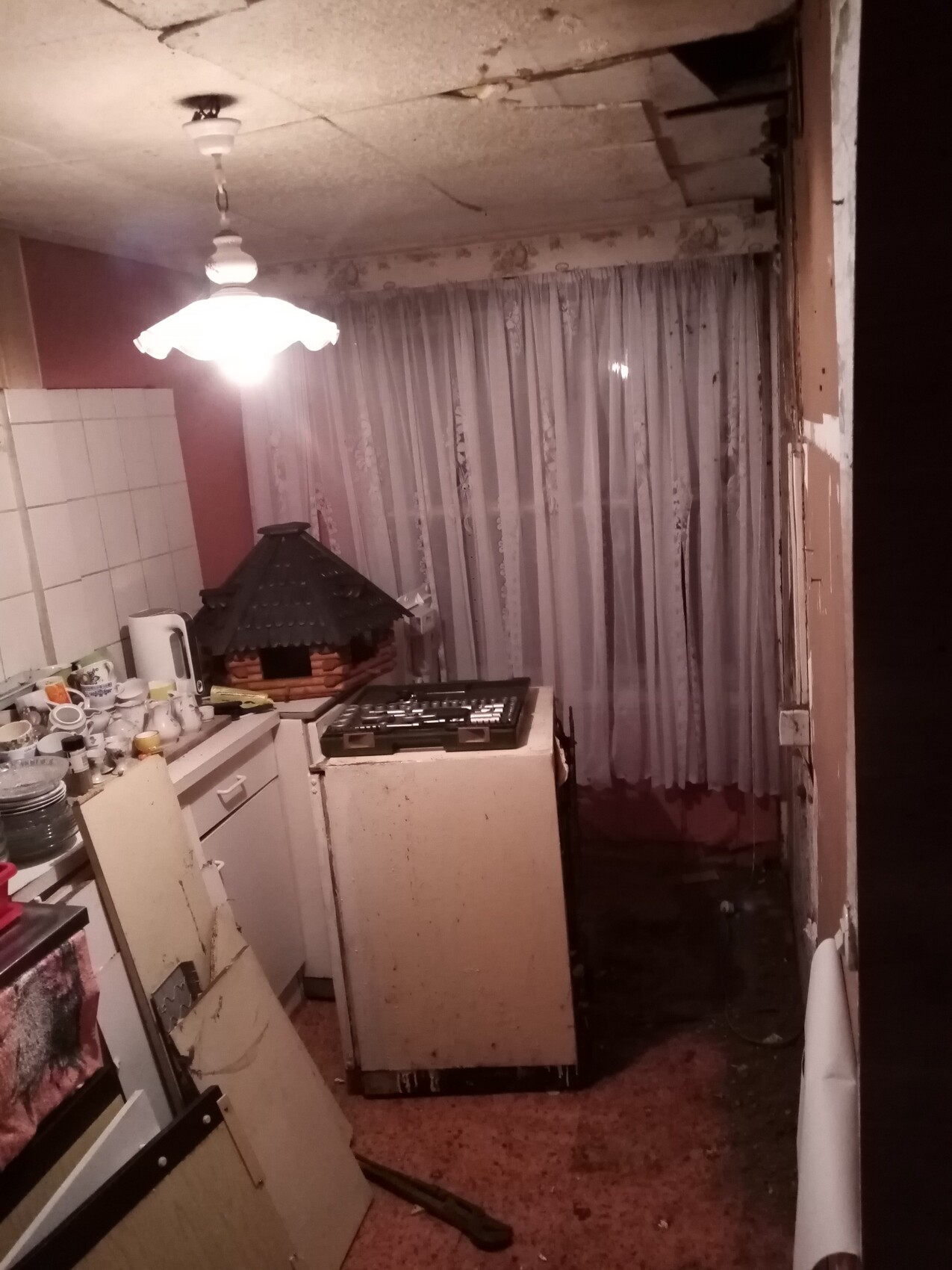 Verlassene Küche: beschädigte Decke, hängende Lampe, chaotische Arbeitsfläche mit Geschirr.