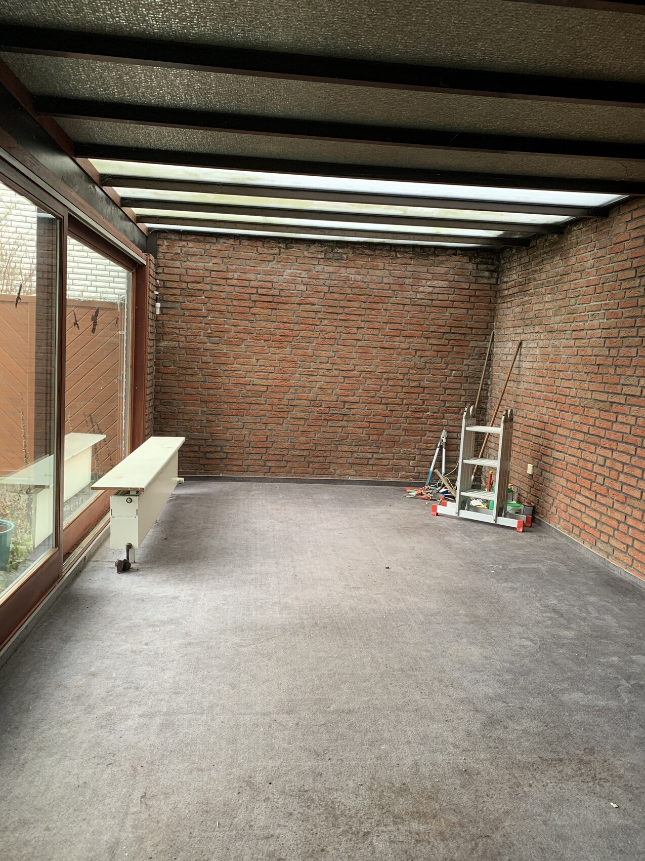 Verglaste Veranda mit Backsteinwänden, Glasdach, linke Fensterfront und Leiter mit Werkzeug rechts.