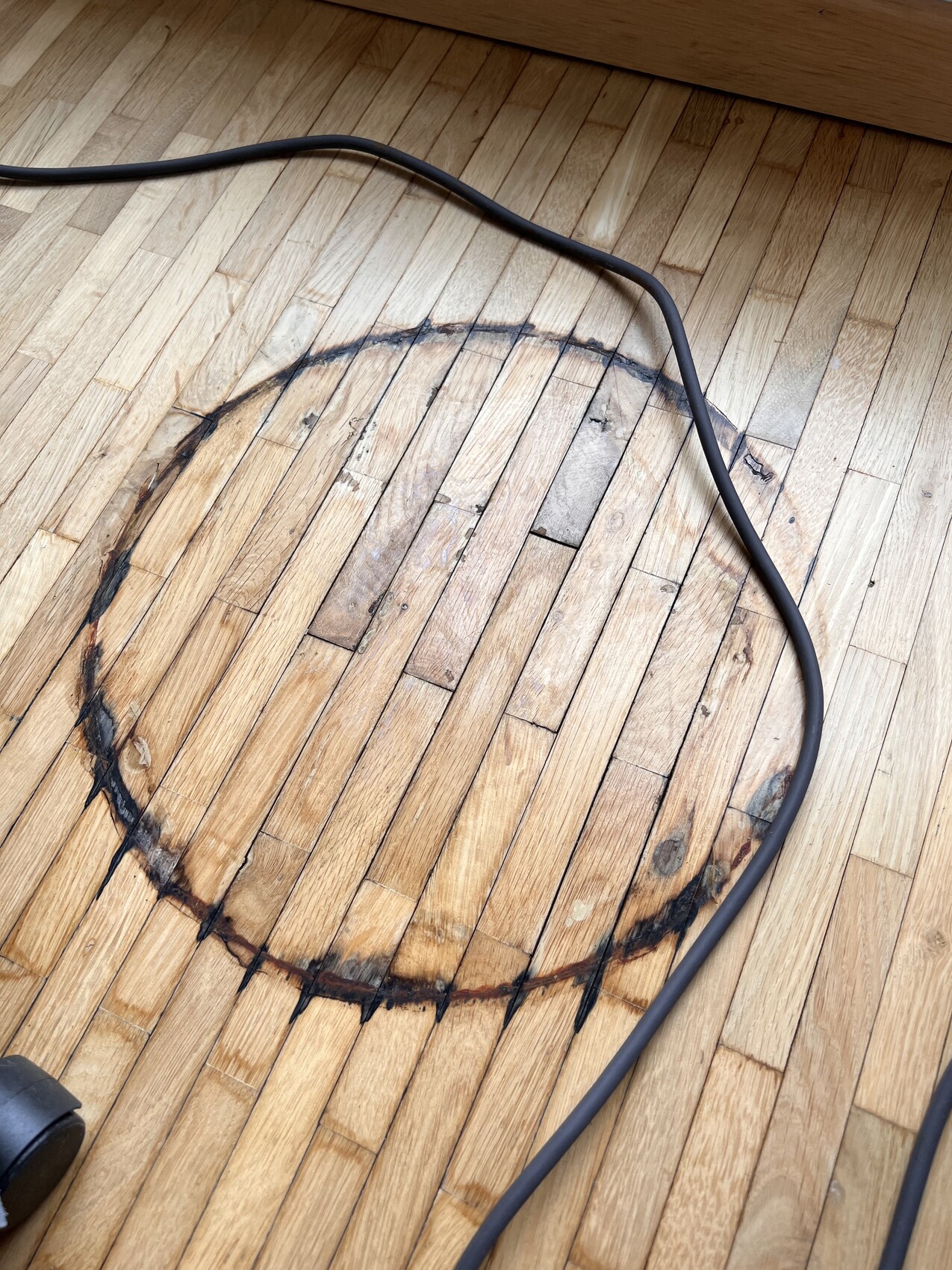 Holzboden mit kreisförmiger Verfärbung, darüber liegt ein schwarzes Kabel.