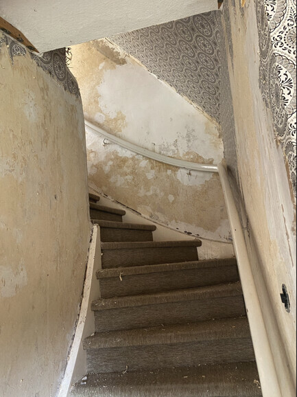 Schmale Treppe in vernachlässigtem Innenraum; abgenutzte Teppichstufen und abgeblätterte Tapete.