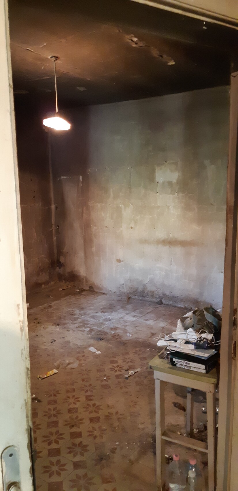 Verlassenes, staubiges Zimmer mit bröckelnden Wänden und einer hängenden Lampe, Tisch voller Schuhe.