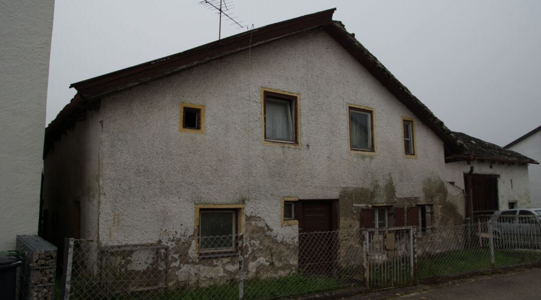 Verfallenes Einfamilienhaus mit beschädigter Putzfassade, alten Fenstern und Zaun.