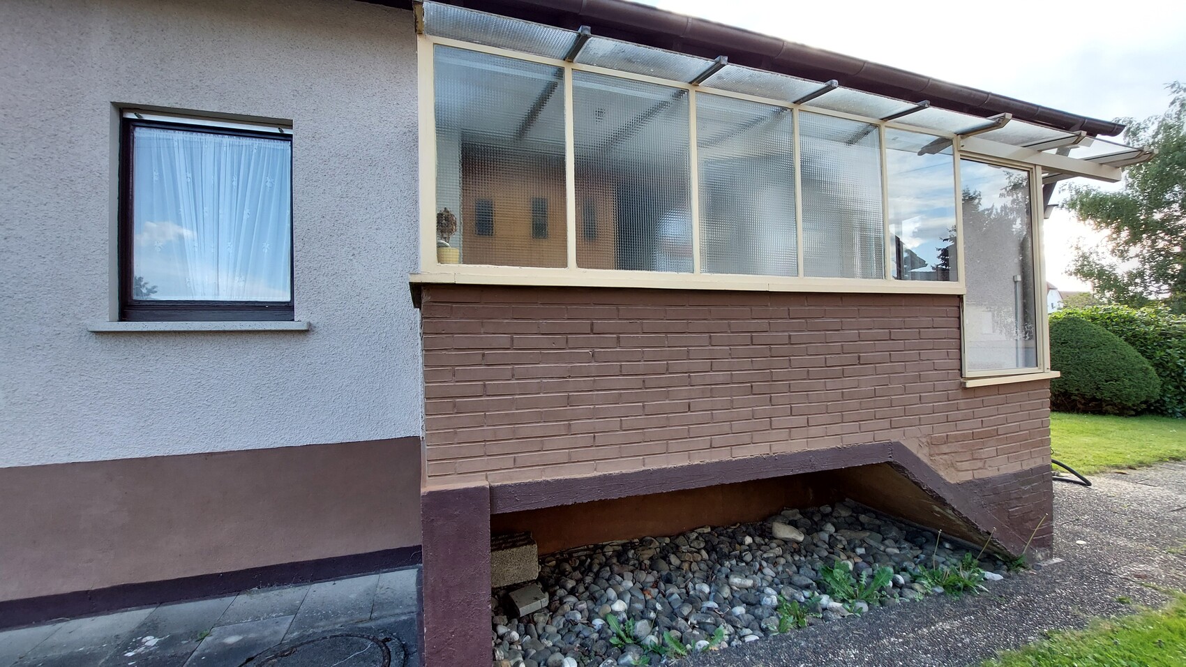 Außenansicht eines Hauses mit verglaster Veranda/Wintergarten, brauner Ziegelwand und Garten.