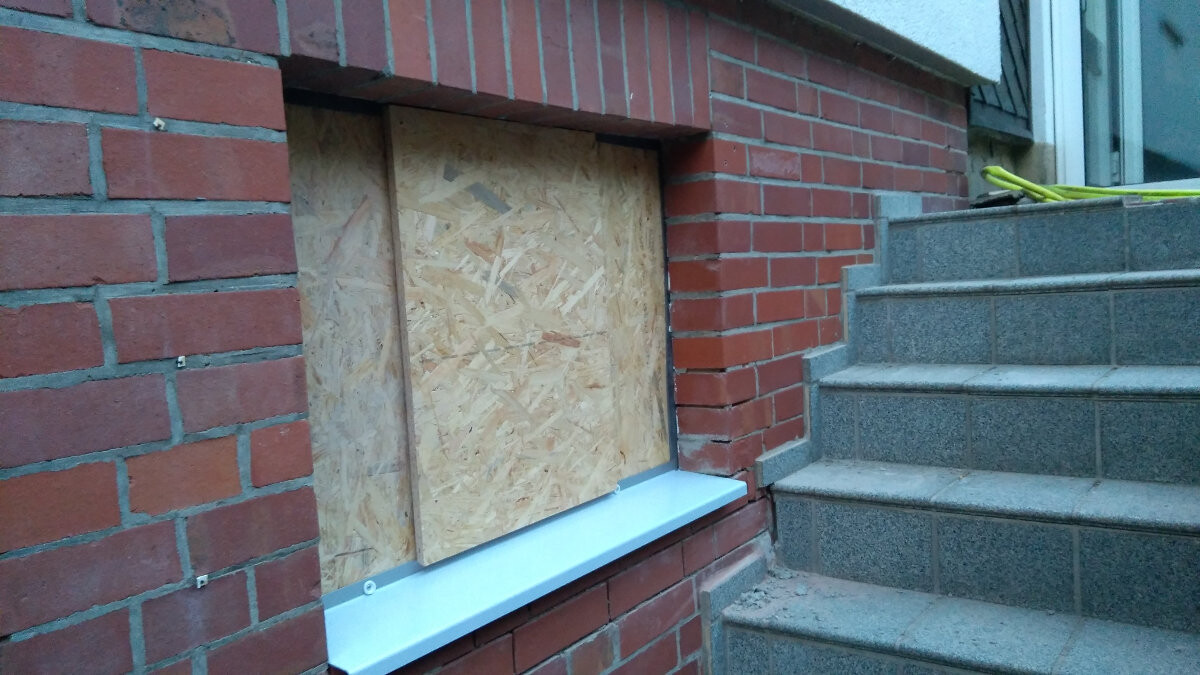 Verschlossenes Kellerfenster hinter OSB-Platte in roter Ziegelwand; rechts Treppen aus Beton.