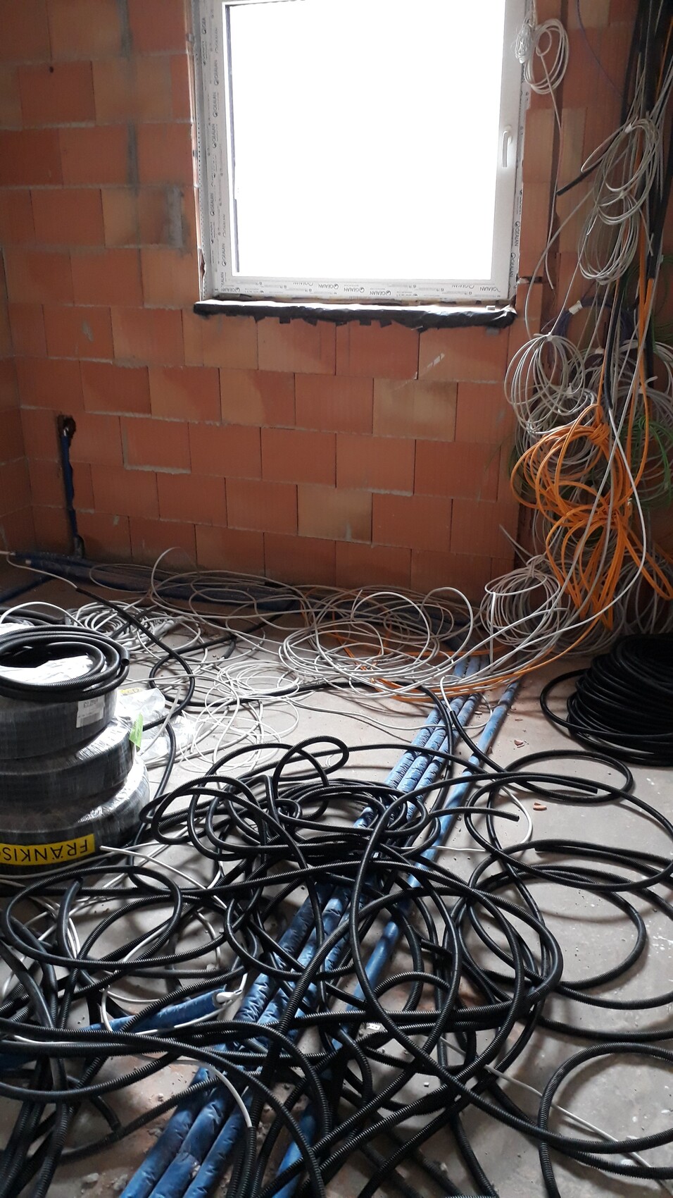 Unfertiges Zimmer mit losen Kabeln, Drähte und Rohren auf dem Boden vor Backsteinwand und Fenster.