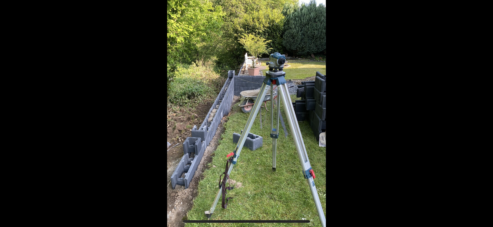 Vermessungs-Tripod mit Theodolit im Garten neben einer Betonstein-Blockmauer und Gartenwerkzeugen.