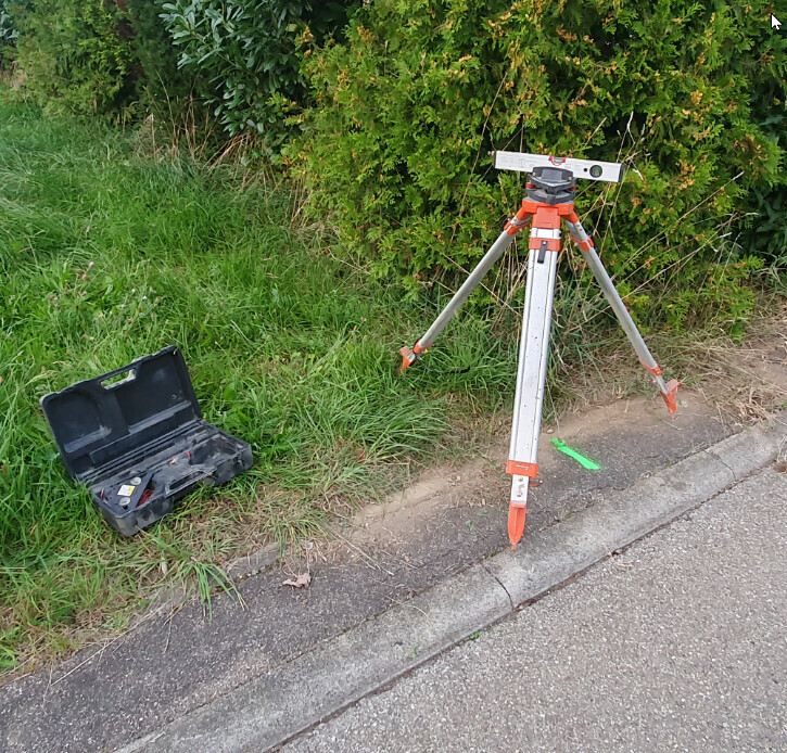 Vermessungsstativ am Straßenrand neben Gras, Koffer mit Vermessungszubehör daneben