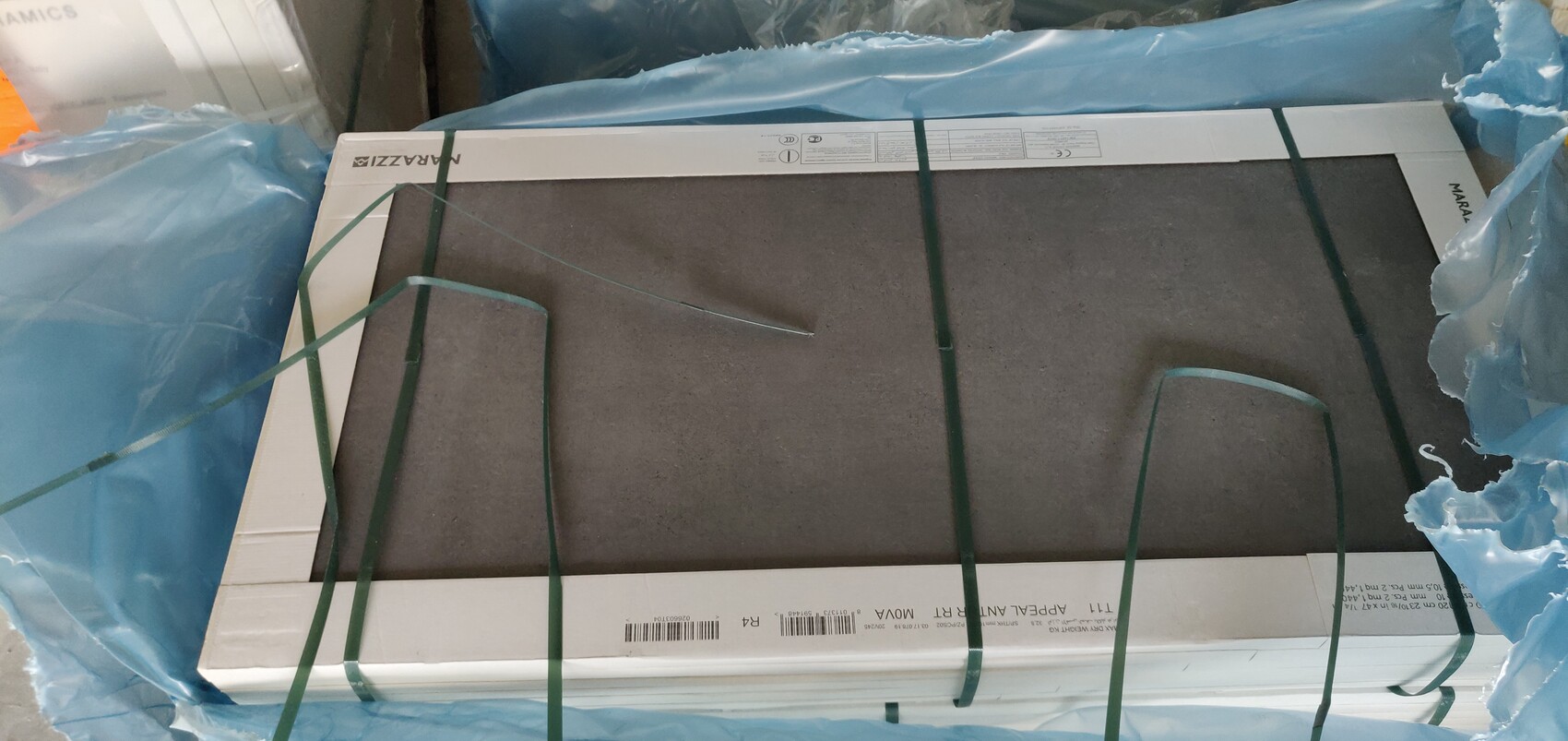 Großformatige, verpackte Fliesenplatte in Karton, umgeben von blauer Folie und grünen Spanngurten.