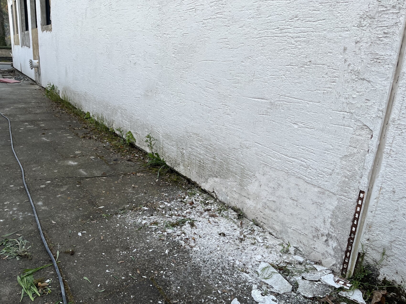 Weiße verputzte Außenwand mit Bodenrissen, Pflanzen am Fundament und Schlauch auf dem Betonweg.