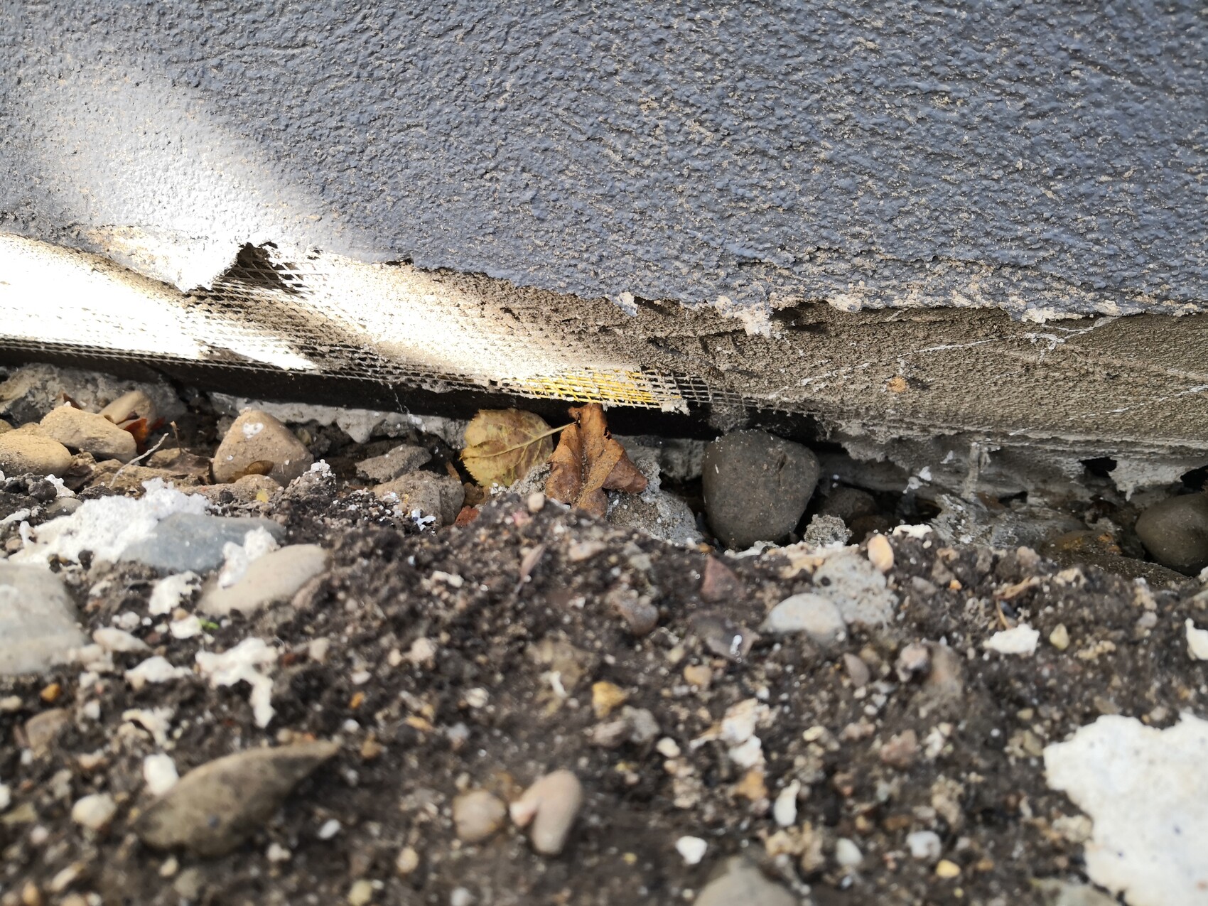 Unterer Wandbereich mit beschädigtem Putz, sichtbarer Netzbewehrung und Kiesboden.