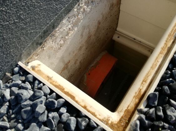 Offene rechteckige Schachtöffnung in einer Wand, Kies um den Rand; roter Gegenstand im Inneren.