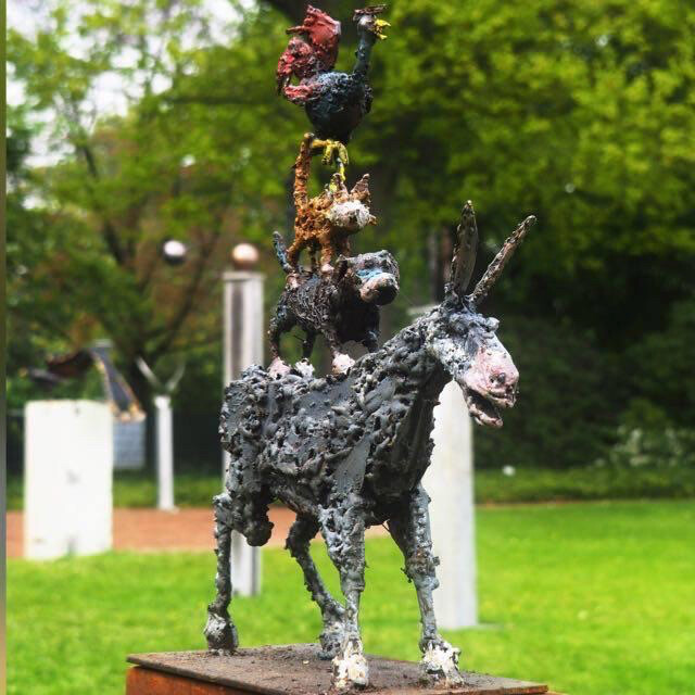 Rostige Metall-Skulptur eines Esels mit mehreren Figuren auf dem Rücken im Park.