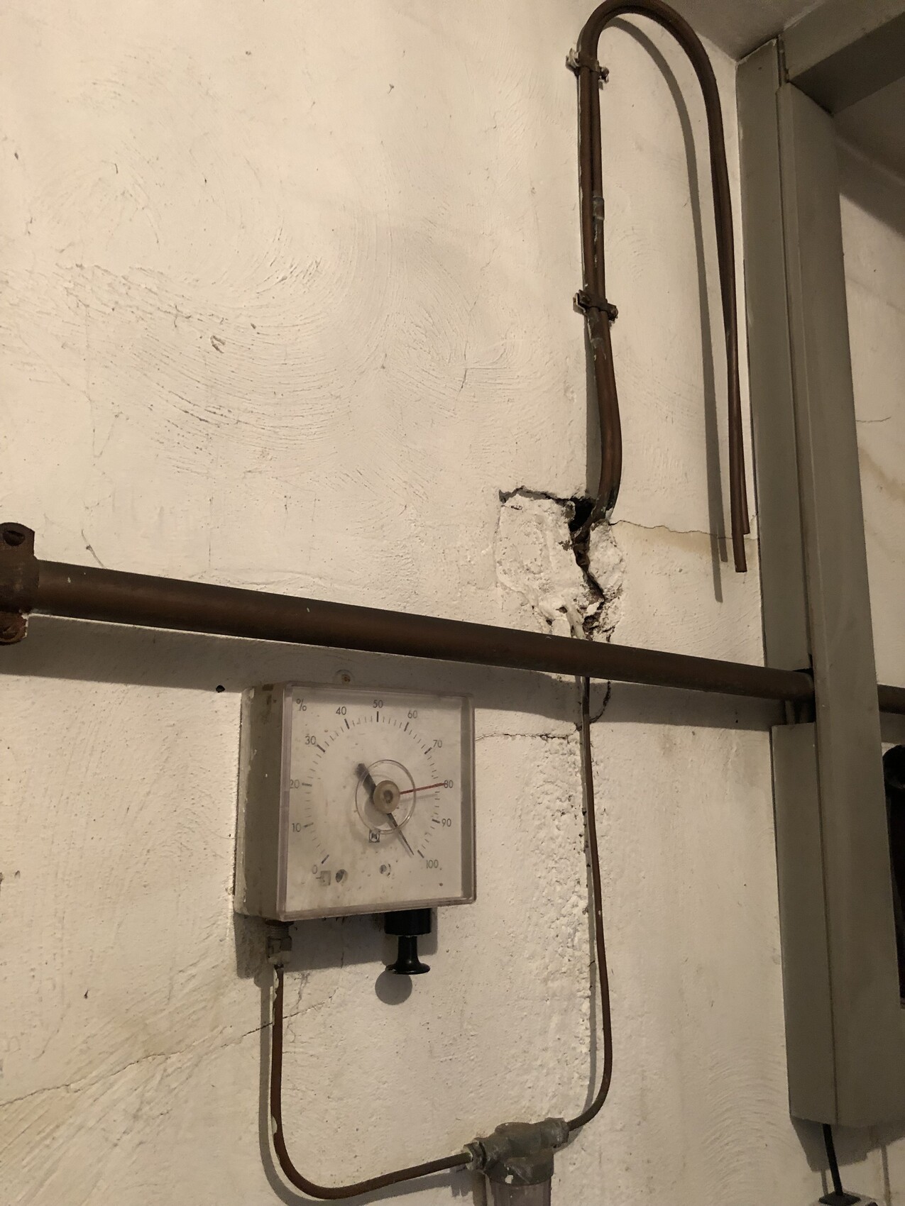 Rostige Metallrohre verlaufen an einer rauen weißen Wand; darunter hängt ein Druckmessgerät.