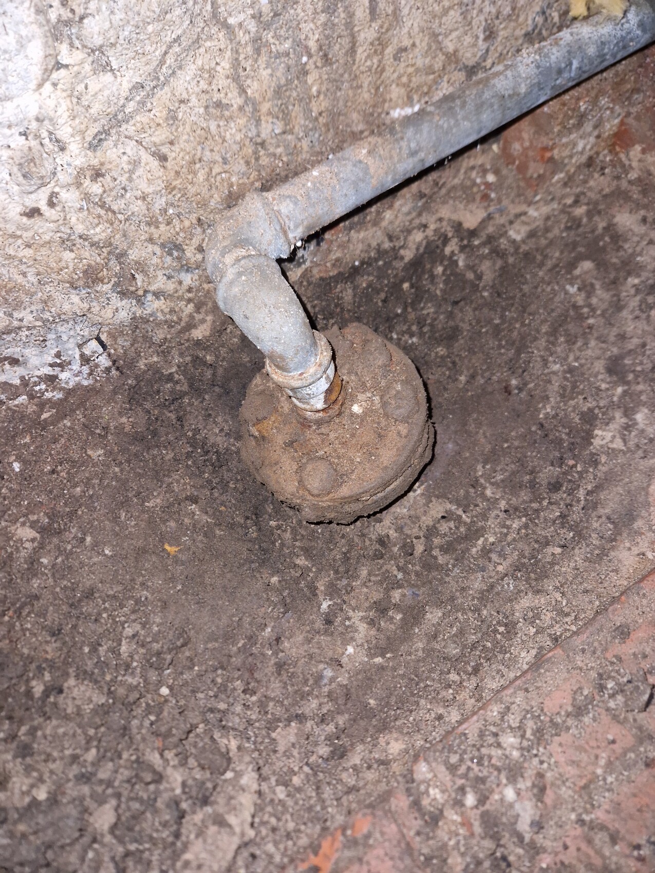 Verrostete Abfluss-Reinigungsöffnung im Erdreich mit gebogenem Rohr darüber.