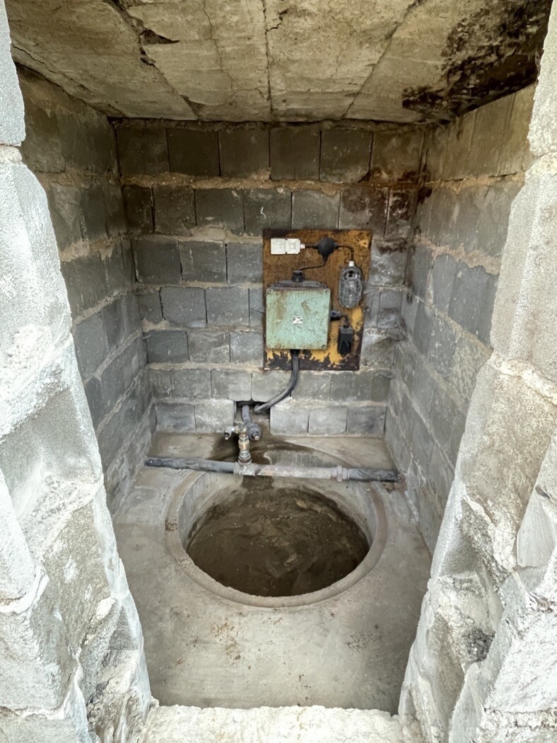 Kleiner Kellerraum mit rundem Bodenloch, Rohrleitungen und rostigem Metallkasten an der Wand