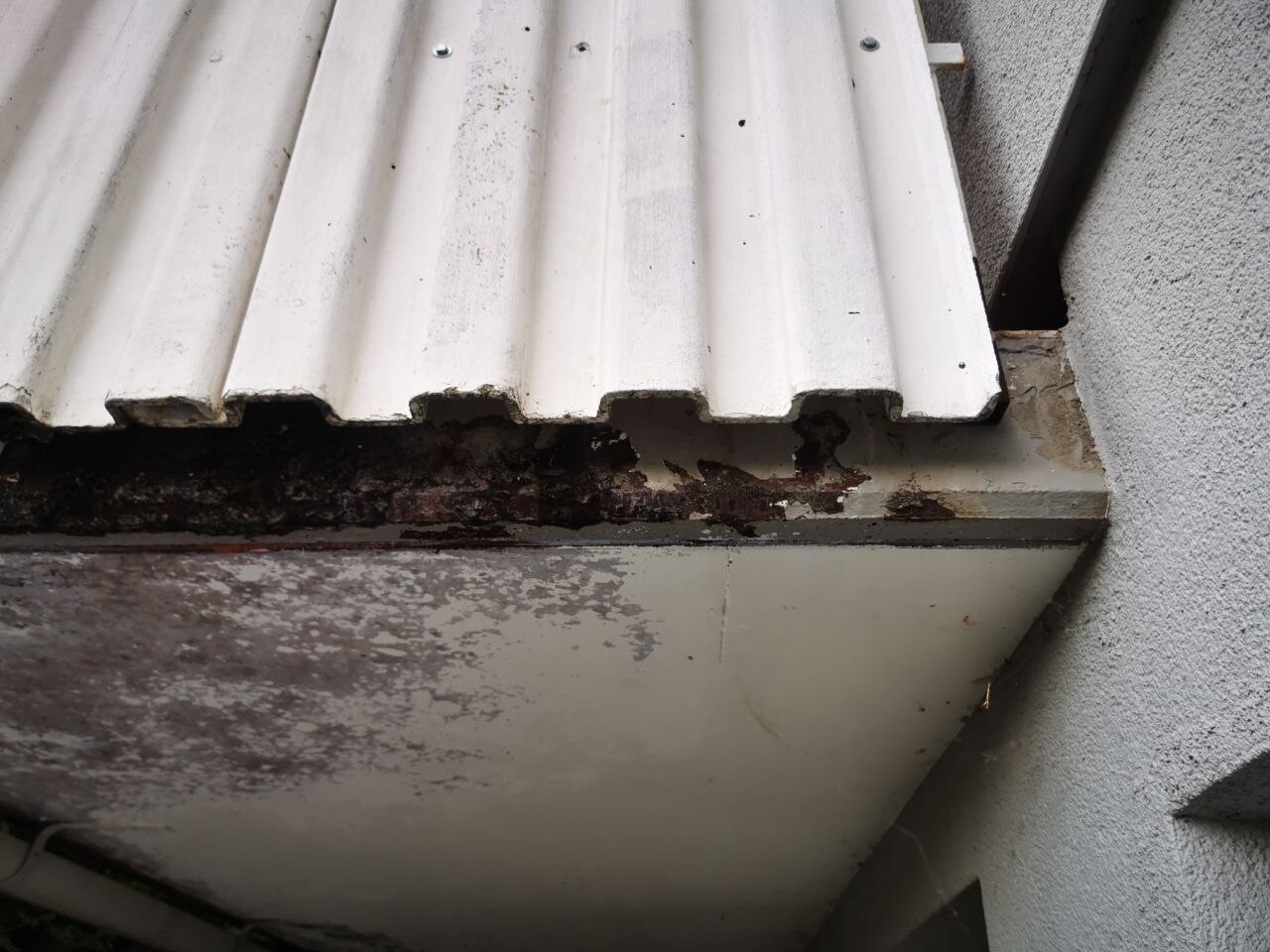 Weißes Wellblechdach mit Rost und Abplatzungen an einer verschmutzten Außenwand.