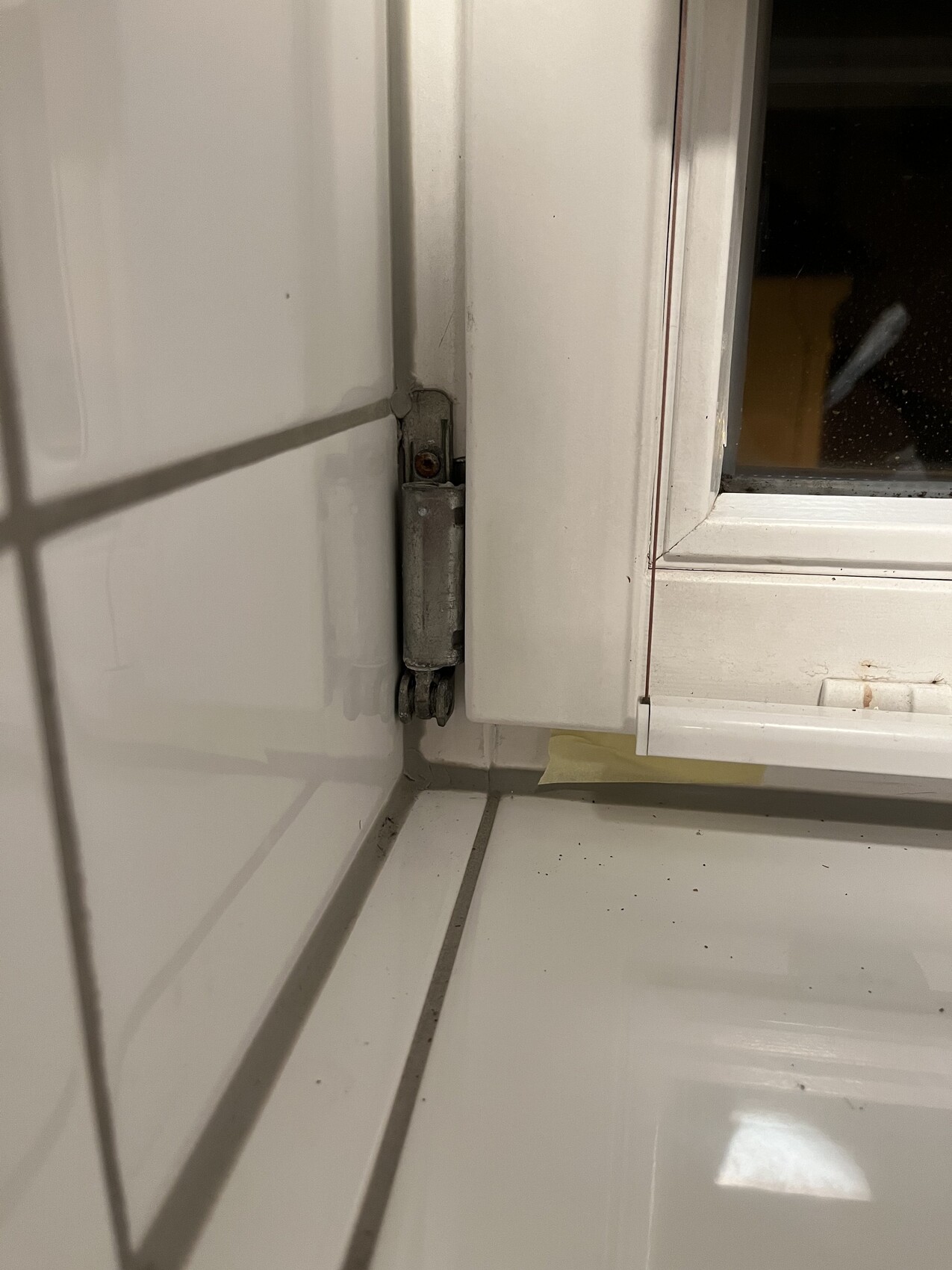 Metall-Scharniere am weißen Fensterrahmen in einer Fliesenwand-Ecke, Staub auf der Fensterbank.
