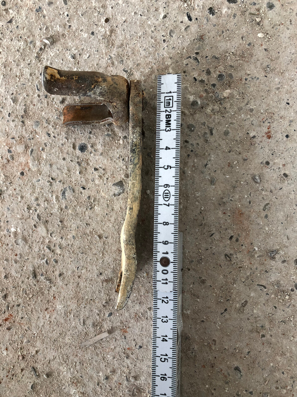 Rostiges Metallwerkzeug mit Haken liegt neben einem Messband auf einem Betonboden.