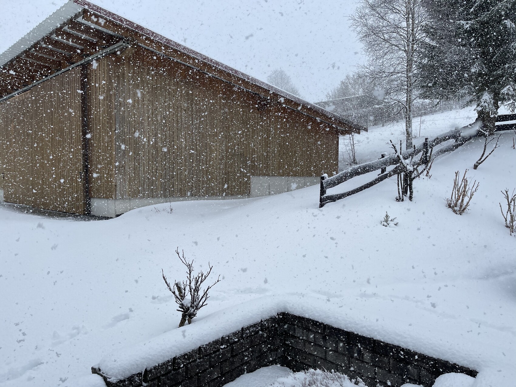 Schneebedeckte Holzhütte mit geneigtem Dach, fallender Schnee, Holzzaun und verschneiter Garten.