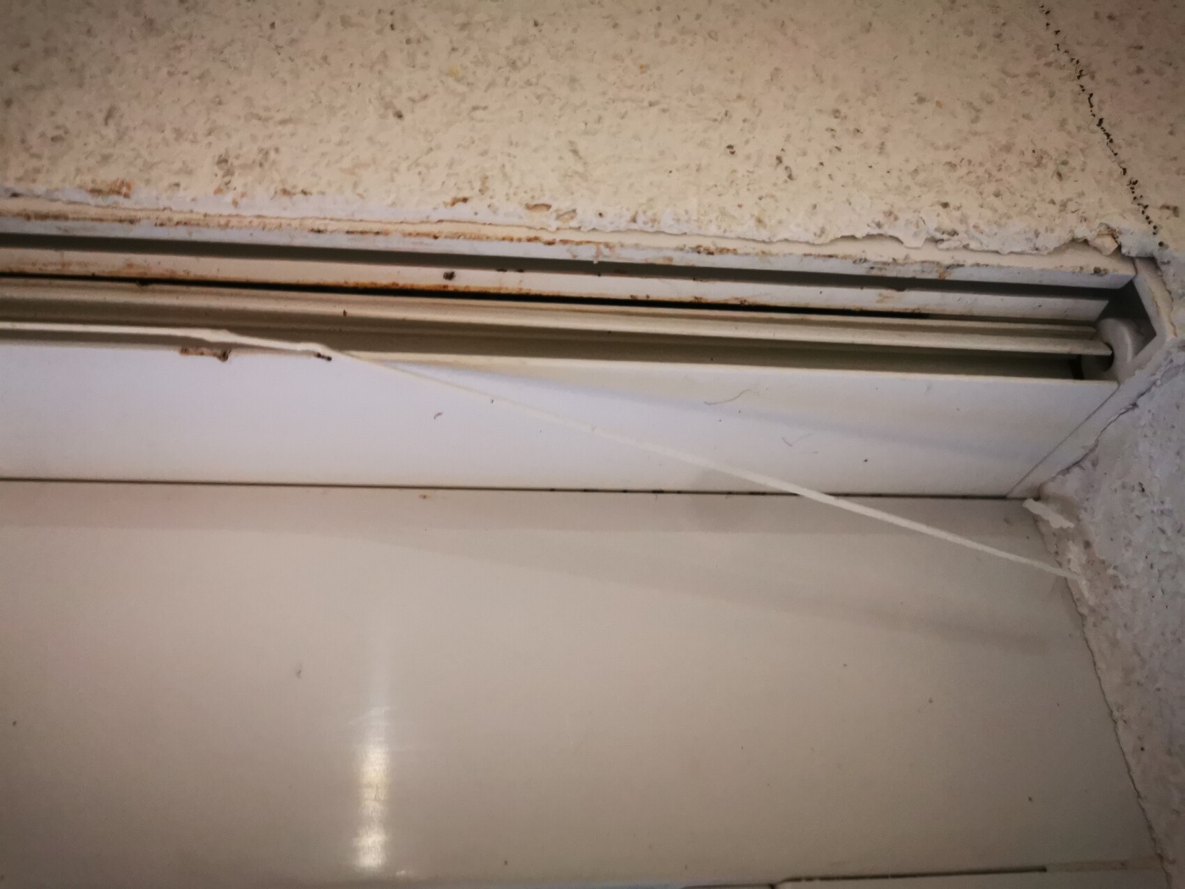 Nahaufnahme einer staubigen Decke mit Rollladen-Schiene und einer diagonalen weißen Schnur.