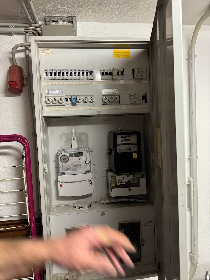 Elektrischer Schaltschrank mit Zählern, Sicherungen und Anzeigen; Hand im Vordergrund.
