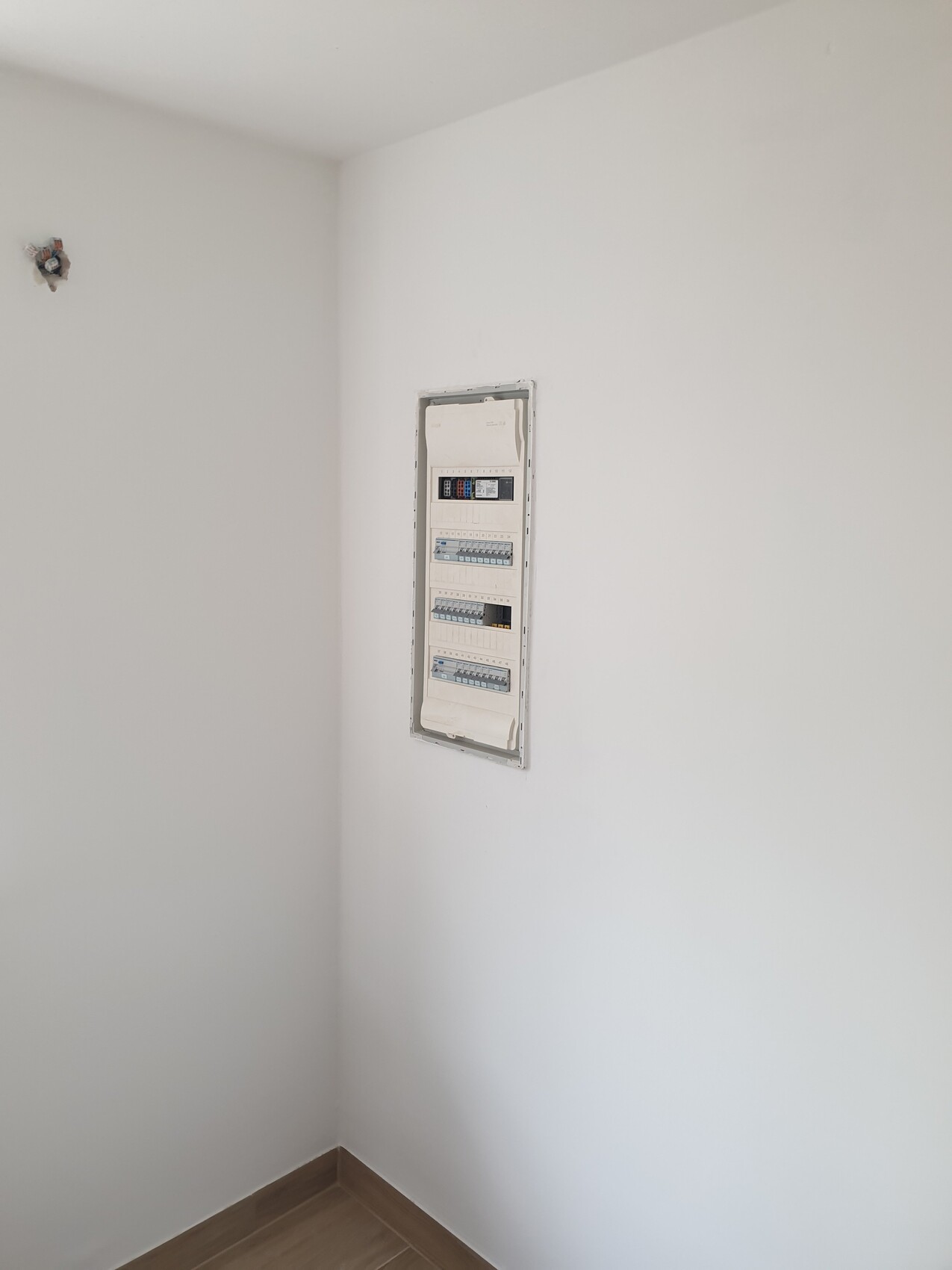 Weiße Wand mit vertikalem Verteilerschrank (Sicherungskasten) in einer Wandnische neben Bodenleiste aus Holz.