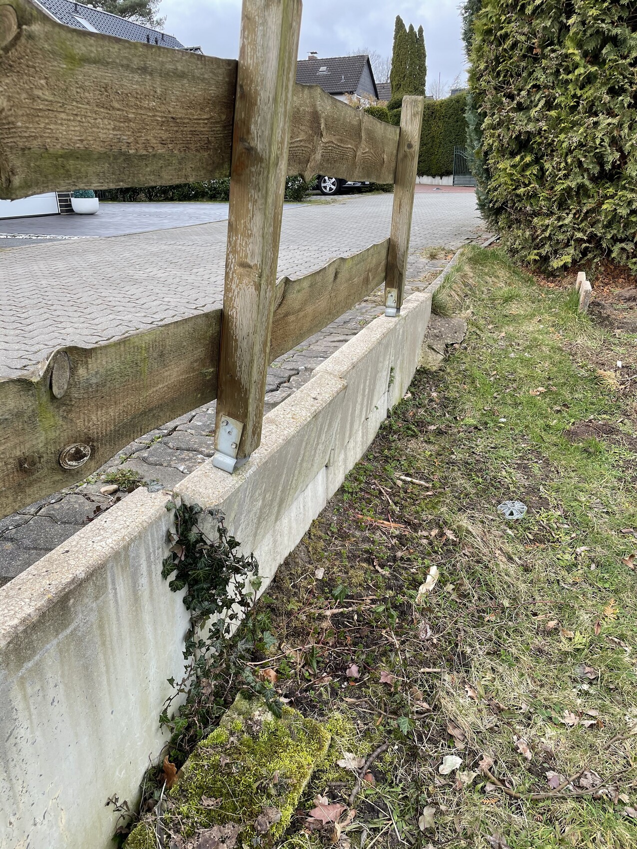 Wettergealterte Holzgeländer auf Betonmauer neben gepflasterter Zufahrt.