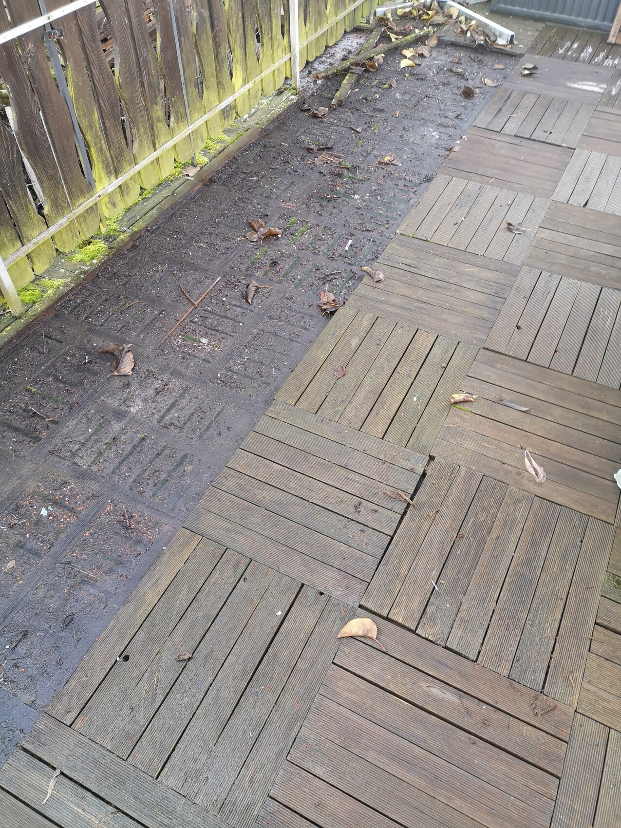 Holzdeck mit rechteckigen Dielen; daneben schmutziger Erdstreifen mit Blättern und Moos am Zaun.