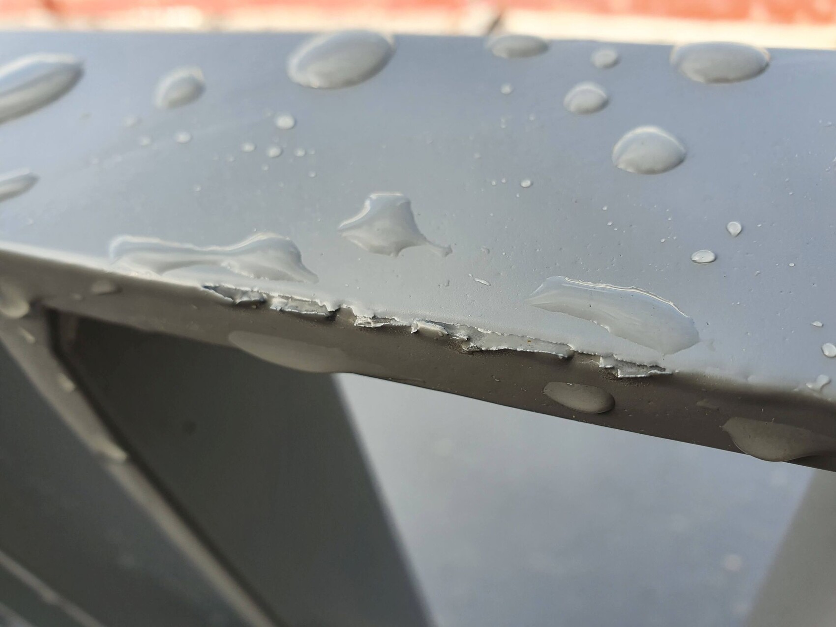 Nahaufnahme einer grauen Metallkante mit Wassertropfen und abgeplatztem Lack.