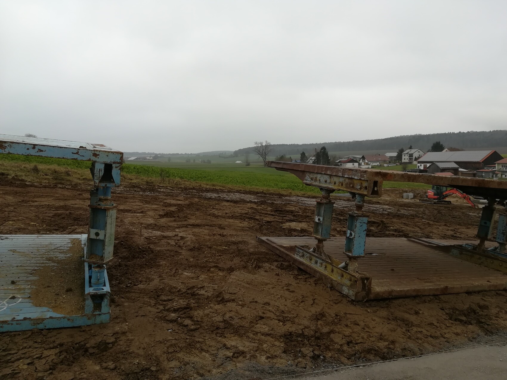Rostige Metallträger auf einer Baustelle im schlammigen Boden; Felder und Häuser im Hintergrund.