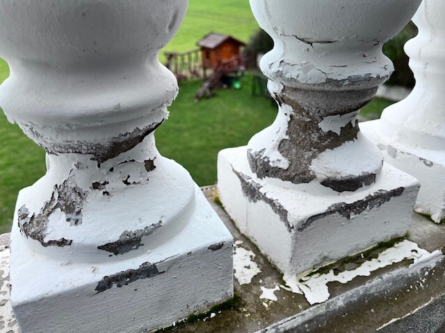 Weiße, abgeplatzte Balustrade im Vordergrund; grüne Wiese und Spielhäuschen im Hintergrund
