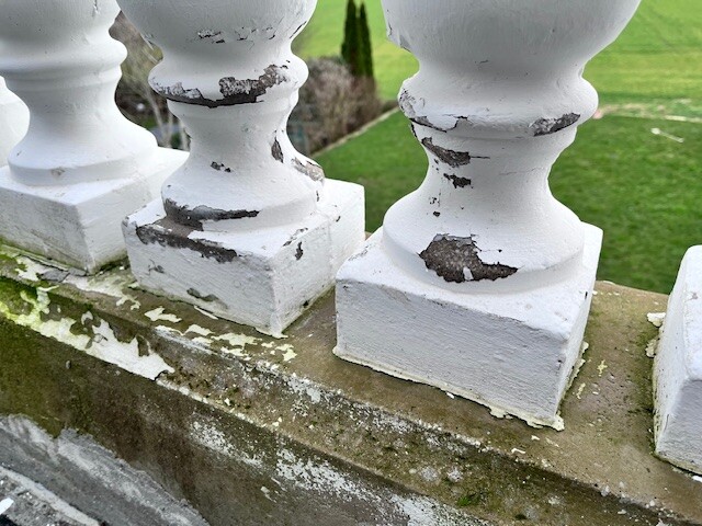 Weiße Balusterpfosten eines Geländers mit abgeplatzter Farbe, Garten im Hintergrund.
