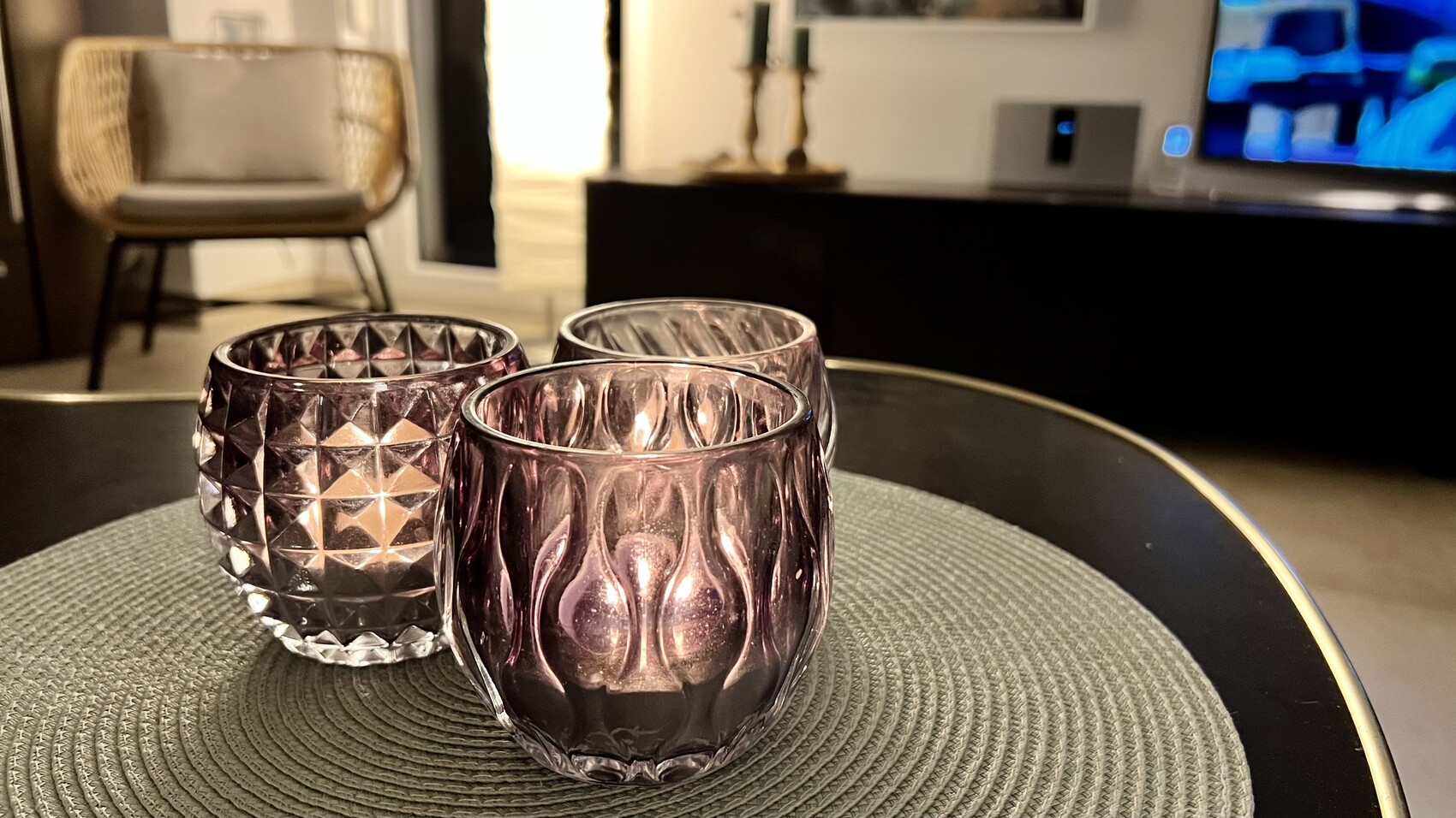 Drei violette Teelichtgläser aus Glas auf runder, gewebter Tischmatte im Wohnzimmer.