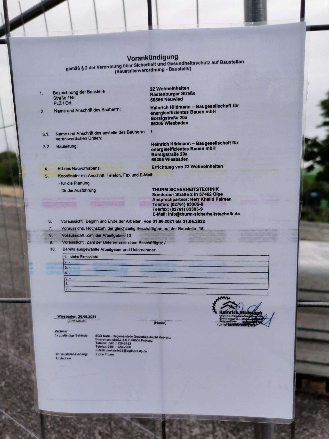 Auf einem Bauzaun befestigtes Vorankündigungsblatt mit Firmenname, Kontakten und Terminen.
