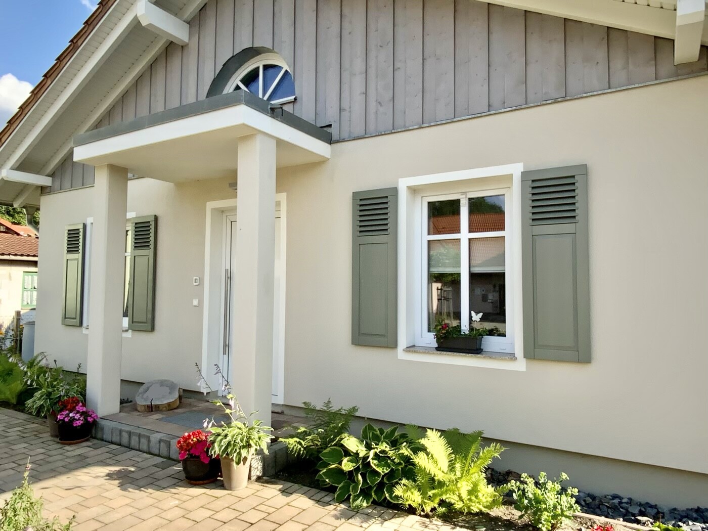 Vordach-Eingang eines Hauses mit zwei Säulen, grauen Fensterläden und Topfpflanzen am Eingang.