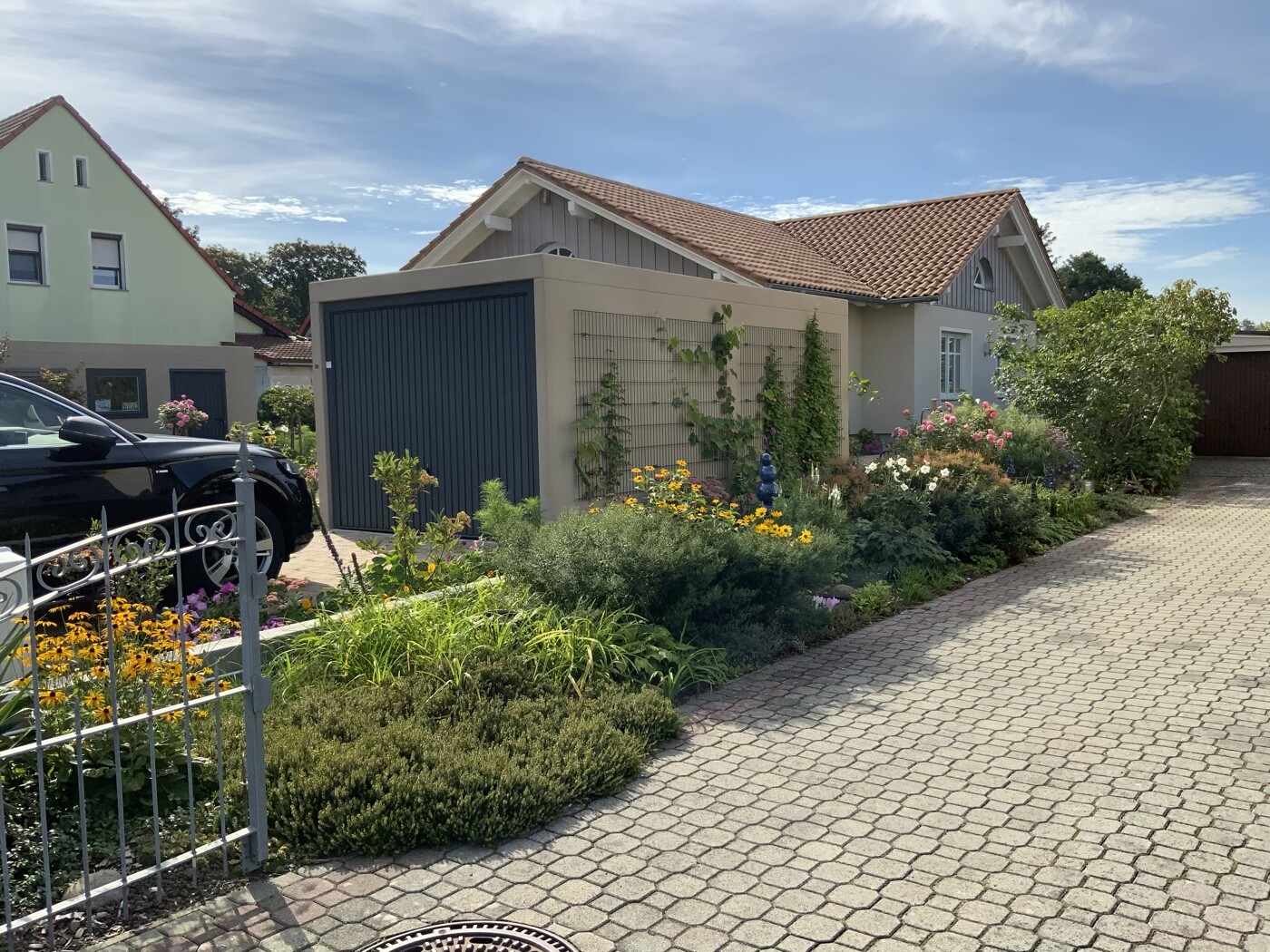 Häuserfront mit Garten, gelbe Blumen, Kletterpflanzen an Garage, gepflasterter Weg, schwarzes Auto.