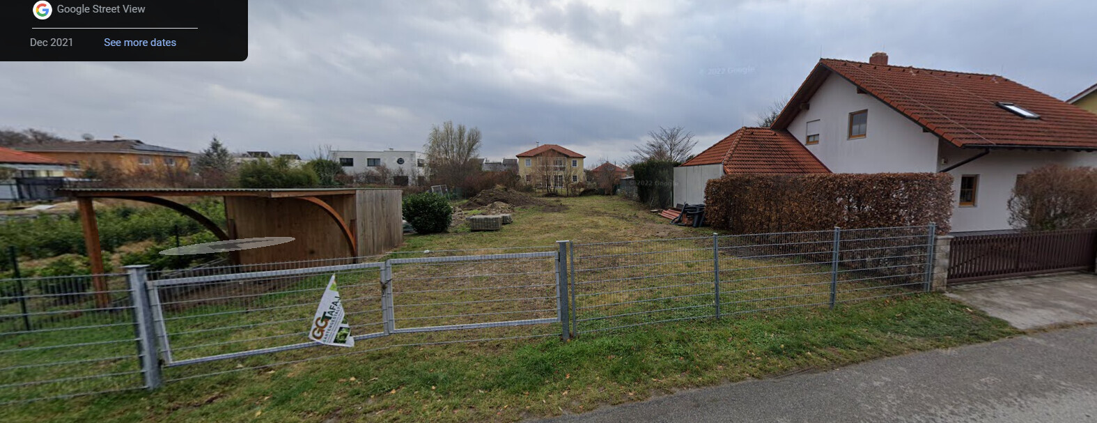 Garten hinter Metallzaun mit Tor, linke Holzkonstruktion, rechts Wohnhaus mit rotem Ziegeldach.