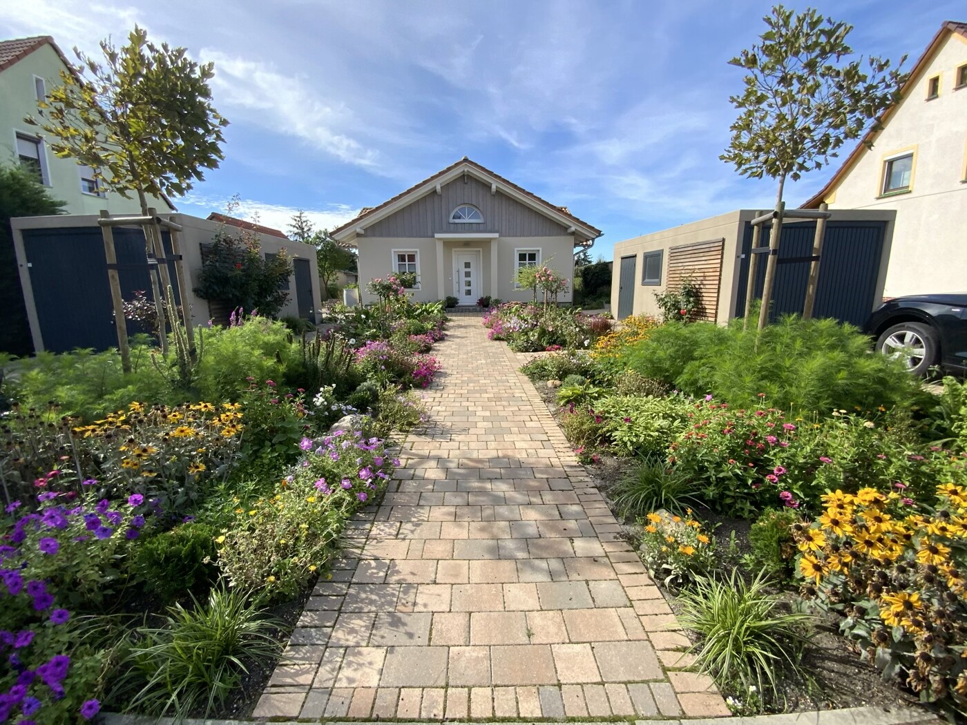 Gepflegter Pflasterweg führt zu einem kleinen Haus, rechts und links bunte Blumenbeete.