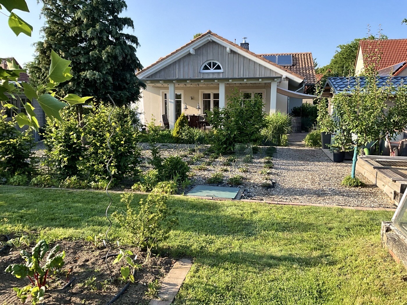 Garten vor einem Einfamilienhaus: Kiesflächen, Bäume, Sträucher und Veranda-Sitzecke.